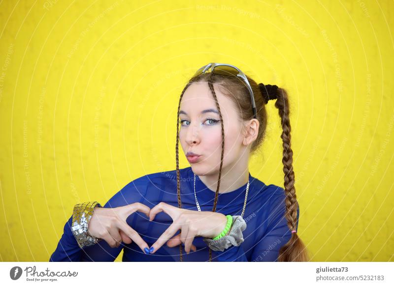 Cool teen girl styled with braids, cute kissing mouth and showing heart with fingers in front of yellow wall 13 - 18 years 18-20 years 17 Youth (Young adults)