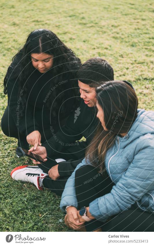 three people of Hispanic-Latino ethnicity, sitting on the ground in the park with smart devices activity atmosphere authentic browsing casual casual. clothes