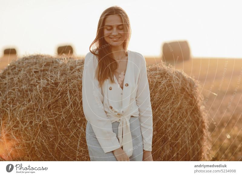 portrait of a smiling beautiful girl with long hair in a jeans skirt. Woman enjoying a walk in a wheat field with hay bales on summer sunny day. woman straw