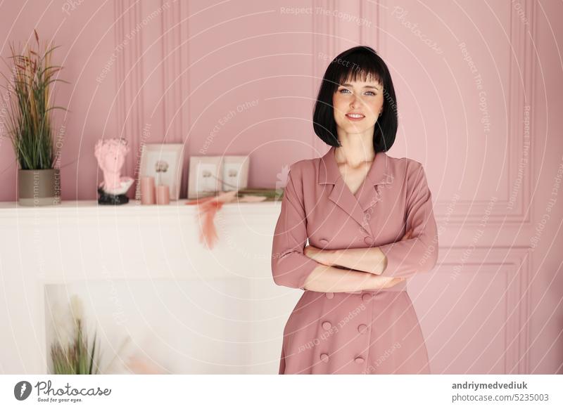 Portrait of attractive smiling brunette with short hair woman folded arms staying alone at stylish interior indoors. Female in soft pink dress is looking in camera