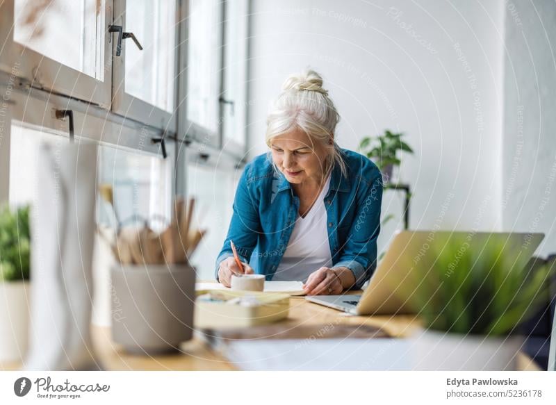 Smiling mature businesswoman writing in notebook while sitting at table in office real people senior indoors loft window home mature adult one person attractive