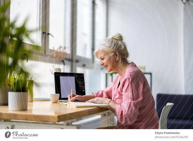 Smiling mature businesswoman writing in notebook while sitting at table in office real people senior indoors loft window home mature adult one person attractive