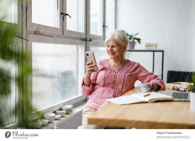 Smiling senior woman using smartphone real people indoors loft window home mature adult one person attractive successful confident beautiful female apartment