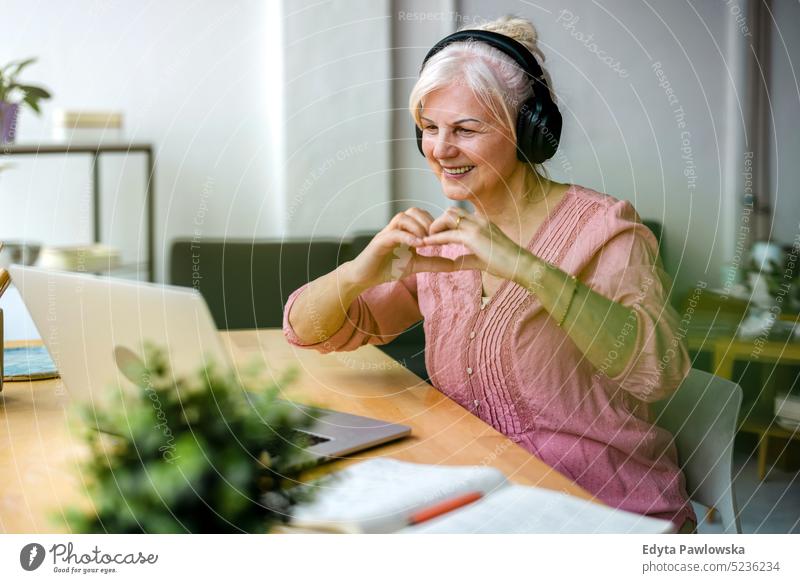 Senior woman forming a heart with her hands during video chat real people senior indoors loft window home mature adult one person attractive successful
