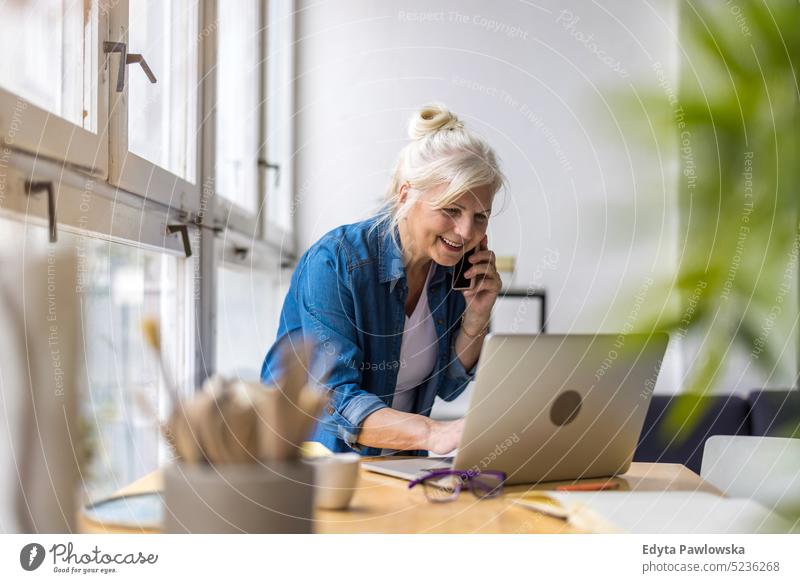 Smiling businesswoman talking on mobile phone while using laptop in office real people senior indoors loft window home mature adult one person attractive