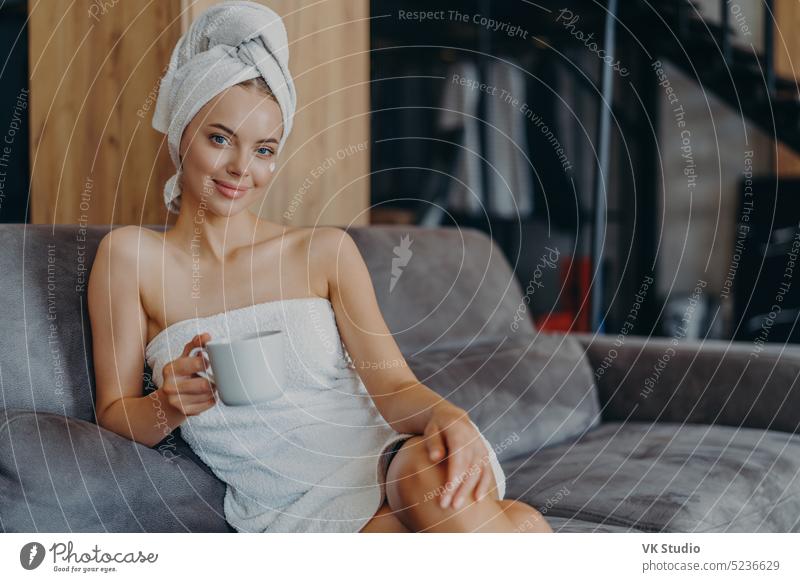 Relaxed beautiful young woman has healthy skin wears soft towel on head and around naked body holds mug of coffee poses at sofa with aromatic beverage enjoys weekends has well groomed complexion