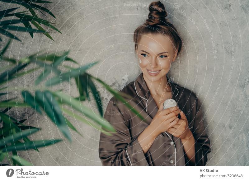 Young caucasian attractive woman in silk brown pajama doing skin care procedure at home beauty young smiling applying face cream cosmetics routine plant green