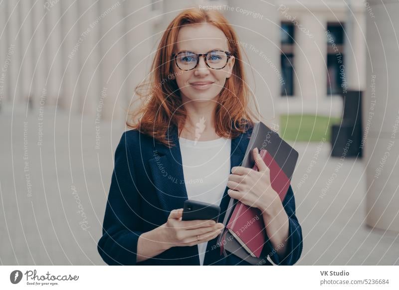Confident beautiful red-haired female business consultant holding modern smartphone and laptop redhead businesswoman eyeglasses mobile posing outdoors city work