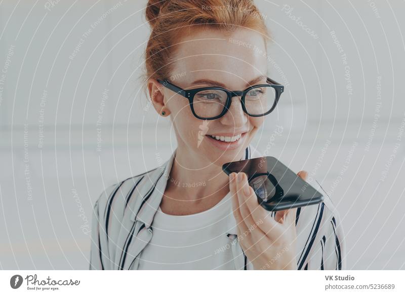 Redhead woman in transparent glasses speaks to virtual assistant holds mobile phone near mouth person face technology voice holding female smartphone talking