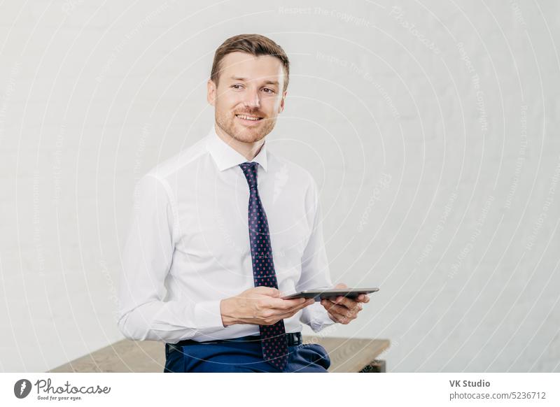 Indoor shot of pleased male owner of prosperous company dressed in formal clothes, holds modern tablet computer, enjoys online communication, looks joyfully aside, has bristle, poses in office