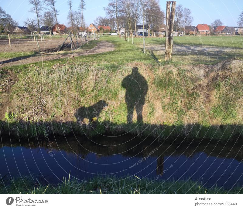 Shadow play on a meadow by the moat showing a dog and a man, in the background a paddock and houses of a village, trees, grass Grass Landscape plants Man Dog