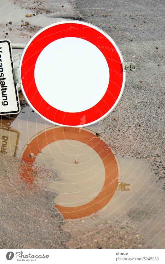 Traffic sign "Durchfahrverbot" with additional sign "Veranstaltung" lie on the asphalt floor and are reflected in a puddle Event Drive-through ban