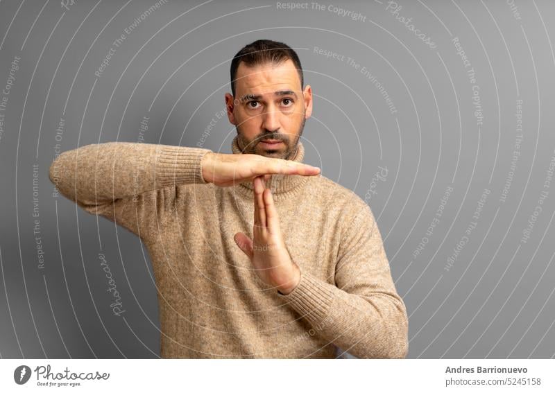 Bearded Hispanic man in his 40s wearing a beige turtleneck making the time-out gesture while looking seriously at the camera, isolated on gray studio background.