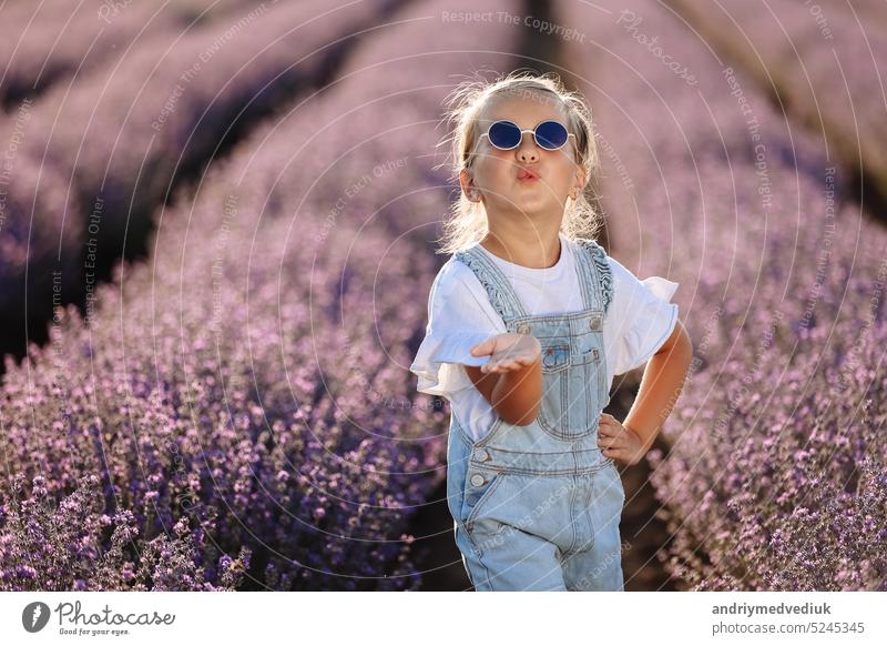 Adorable happy child girl is having fun and blows air kiss at camera in lavender field on summer warm day. Hyperactive little kid in sunglasses on nature. International Children's Day, Valentines Day