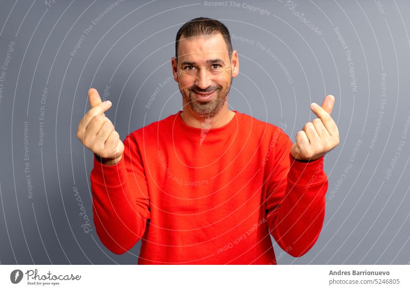 Bearded hispanic man in his 40s wearing a red sweater making the money gesture rubbing his fingers with a greedy face, isolated over gray background. smiling