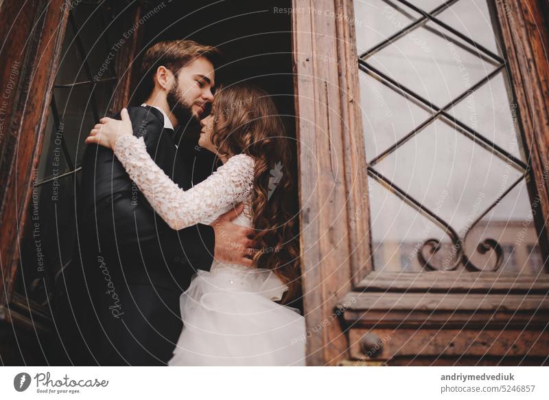 Beautiful newlyweds hugging near the ancient door. Wedding portrait of a stylish groom and a young bride near old house in in a European town embrace beautiful