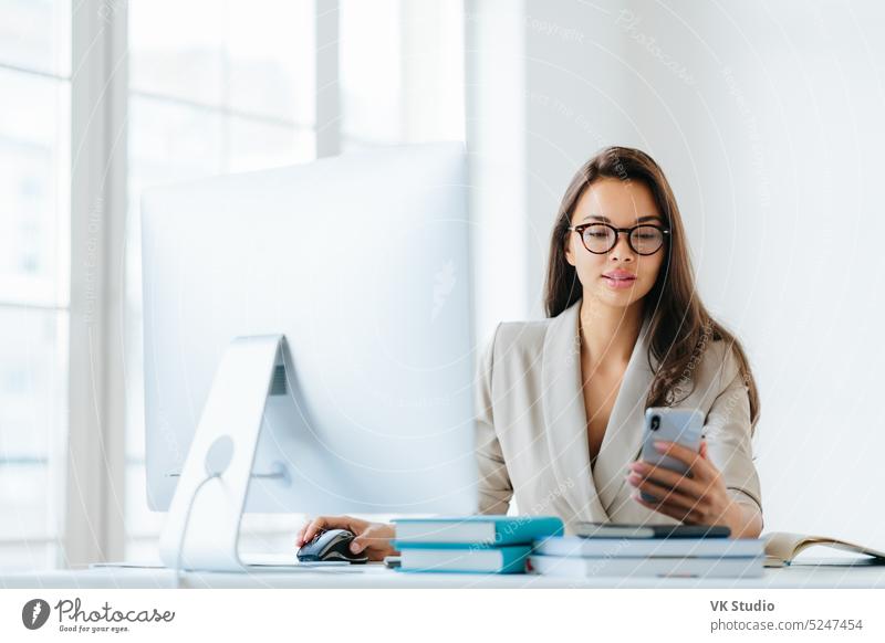 Elegant woman office worker uses mobile phone and computer at one time, sends messages and chats with clients online, monitors news from networks, sits at desktop with pile of notepads and books