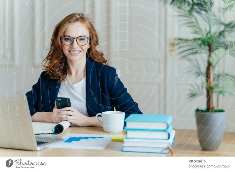 Photo of red haired smiling young woman installs app on mobile phone, checks newsfeed, works on laptop computer remotely, poses in coworking space with cup of drink, books and paper documents
