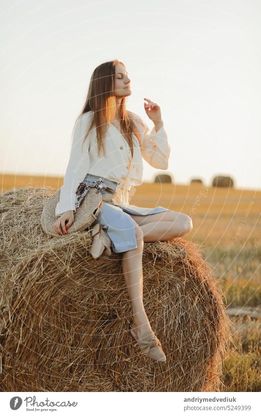 Young woman with long hair, wearing jeans skirt, light shirt and straw bag in hand, sitting on bale on field in summer. Female portrait in natural rural scene. Environmental eco tourism concept.