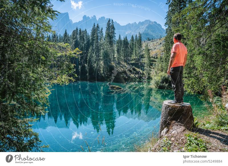 An adult man admiring the wonderful mountain scenery of the turquoise water of the moraine Carezza Lake lake and silhouette of Latemar mountain group peaks in the background.Dolomites, Italy
