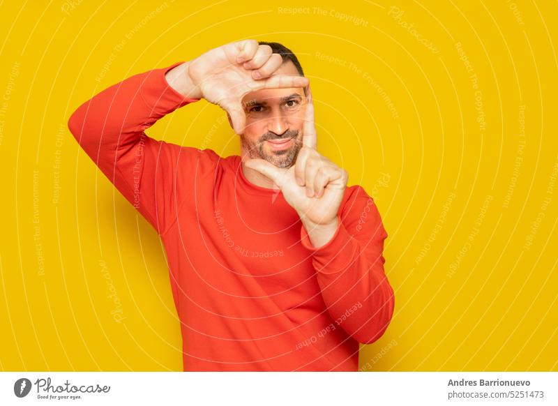 Bearded Hispanic man in his 40s wearing a red sweater making a frame with his fingers, isolated on yellow background. Creativity concept. beard hand gesture