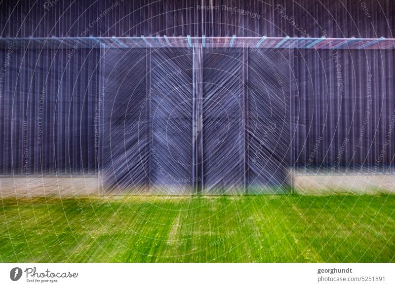 A double sliding gate made of stained wooden slats on a barn. Black and brown with concrete colored base and a green lawn in front of it. The image has been zoomed in and motion blur can be seen.