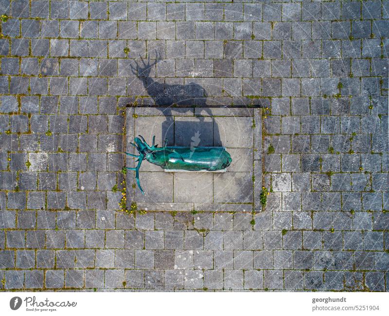 Bronze deer Blankenberg | aerial view of a statue of a deer above a pavement with square stones. The statue is made of bronze and oxidized turquoise. It casts a shadow through which the figure can be seen better.