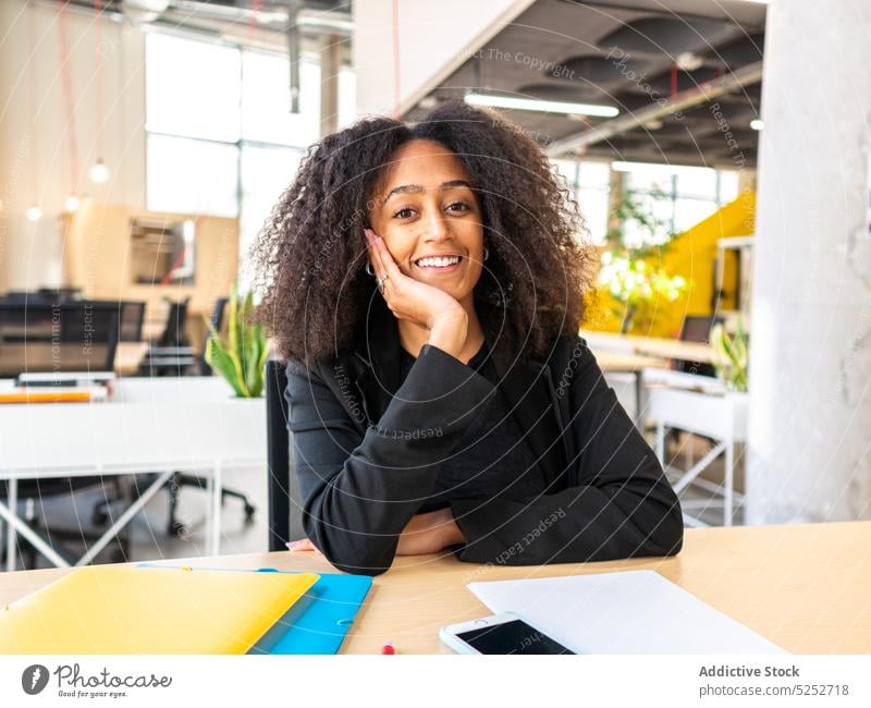 Dreamy black female employee sitting in office woman lean on hand dreamy table smartphone company workspace occupation contemplate smile young african american