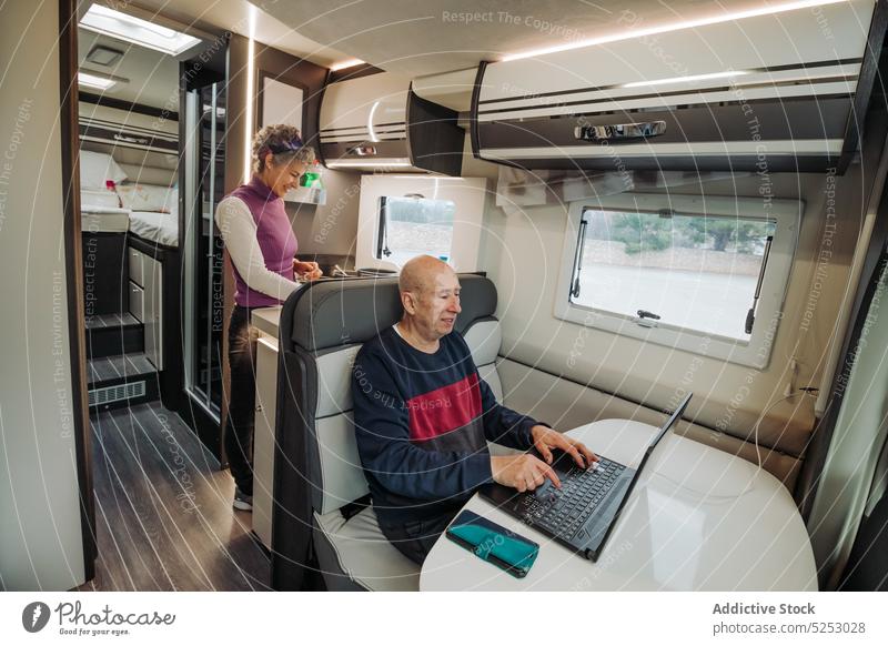 Happy woman cooking food while man using laptop in camper van couple typing road trip journey positive mature aged modern travel pleasure dinner netbook