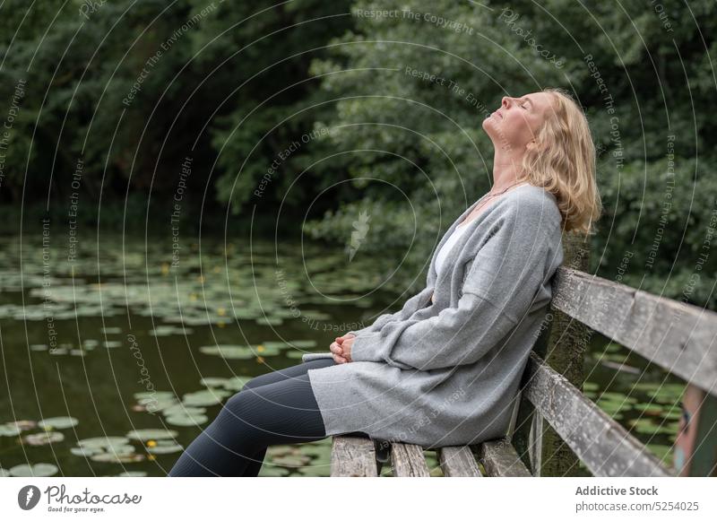 Relaxed woman sitting on bench near lake rest lotus leaf tree relax countryside pleasure water dreamy contemplate middle age female cardigan pleasant enjoy calm