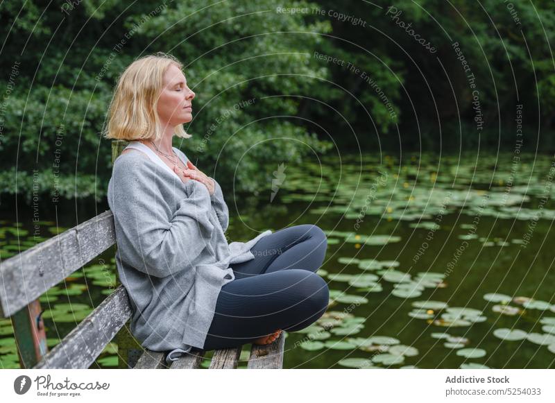 Calm woman sitting in lotus pose near pond padmasana meditate bench lotus leaf eyes closed recreation middle age female enjoy spirit lake yoga stress relief