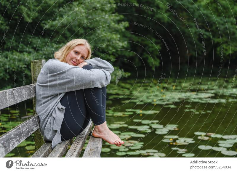 Calm woman embracing knees sitting on bench near pond rest lotus leaf tree park recreation countryside contemplate middle age female enjoy chill lake weekend