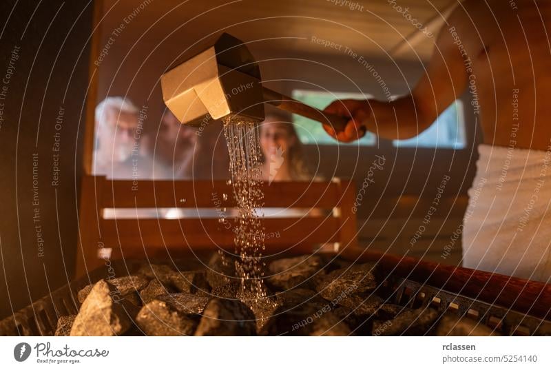 man pouring water onto hot stones with rain spoon in sauna room with a group of people. Steam an water on the stones, spa and wellness concept, relax in hot finnish sauna.