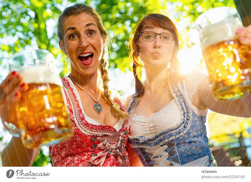 Girlfriends looking and screaming, kissing at camera say cheers or clinking glasses with mug of beer in Bavarian beer garden or oktoberfest amazed party woman