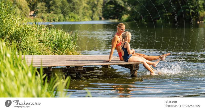 mother and daughter bathing and splashing water with their foot at jetty on a lake in summer kid child reed vacation swimwear bikini bavaria germany