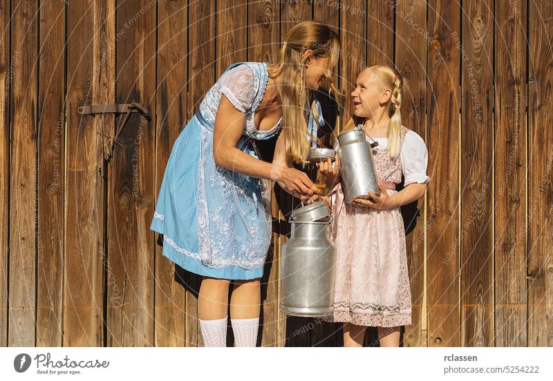 Mother and daughter holding milk cans in traditional Bavarian Tracht at a farm in Bavaria on a wood background. Oktoberfest concept image braid girl child