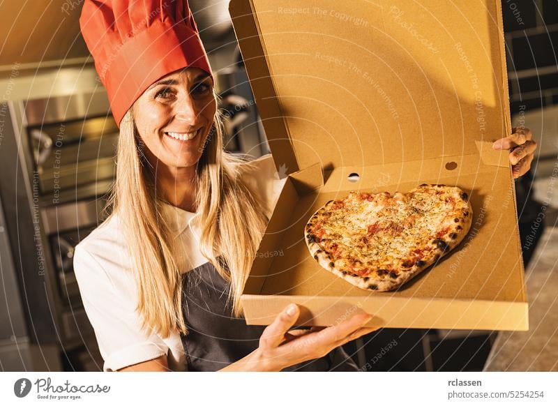 female pizza baker holding a fresh cheese pizza in a heart shape from the stone oven in a box for delivery at italian pizzeria smile valentine love tasty cheesy