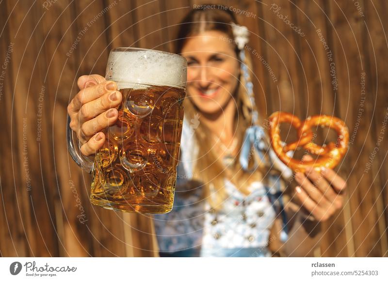 Happy woman toasting with huge mug of beer in traditional Bavarian Tracht, holding pretzel in other hand in Bavarian beer garden or oktoberfest gold party