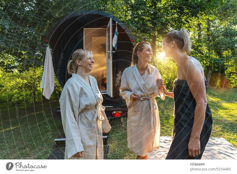 Three girlfriends talking and standing in front of a wooden barrel sauna at Summer in norway. Are relaxed, laughing  and enjoy the holiday while relaxing in the finnish sauna cabin.