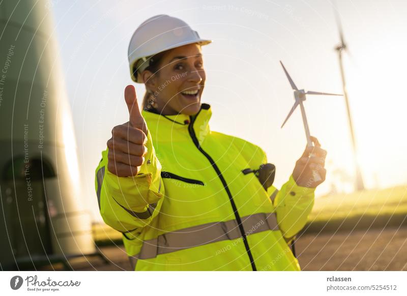 female engineer holding wind turbine model and thumbs up at a windfarm. New Energy concept image safety jacket helmet woman hardhat technician people business
