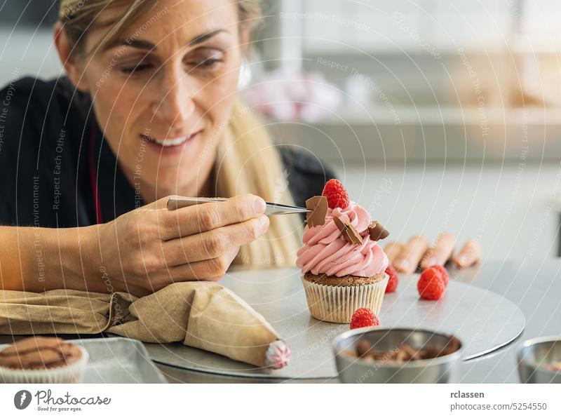 Happy women in pastry bakery as confectioner draped a piece of chocolate on a muffin topping with tweezers icing bag pink raspberry cake cupcake birthday sugar
