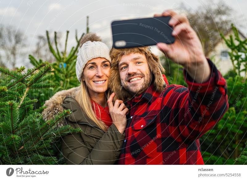 Woman and man holding smartphone and smiling to take a selfie on christmas tree market having fun photo camera pine tree woman people love winter girl sale