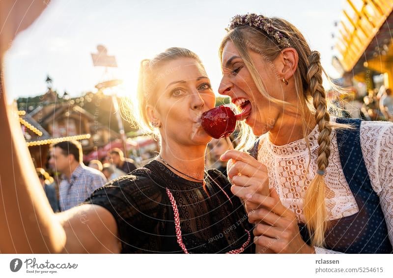 Two friends making selfie wearing dirndl and holding candy apples at a Bavarian fair or oktoberfest or duld in national costume or Dirndl photo smartphome sweet