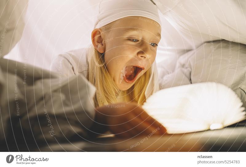 astonished young child reading book under blanket at night. Magic light coming out of the book. childhood girl flashlight kids family home education happy cute