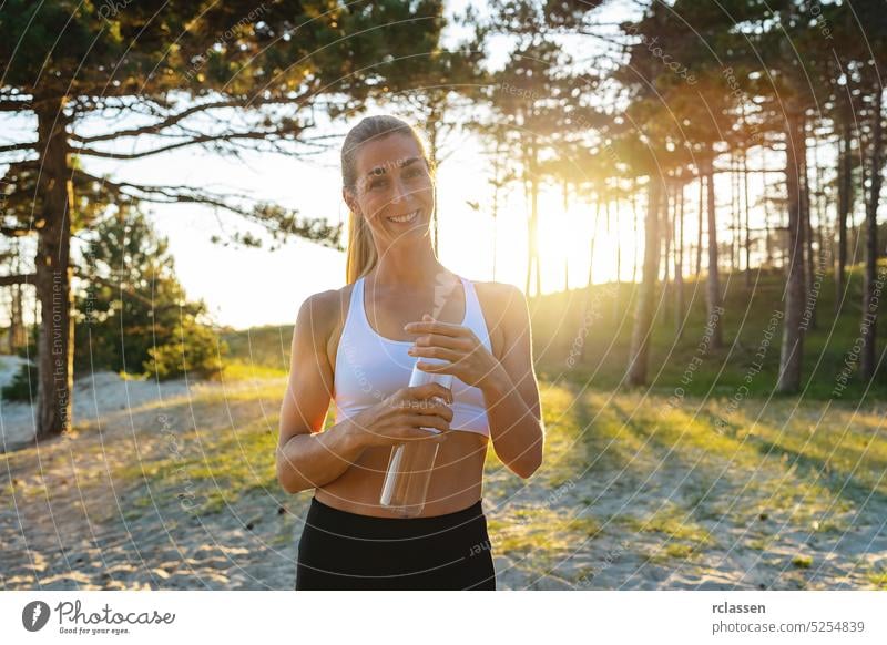 happy woman athlete takes a break at the beach forest, holding water bottle and looking at camera on a hot day. sunrise drink attractive woman exercise sport