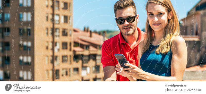 Young couple with mobile looking at camera in rooftop on a summer day smiling friends portrait looking camera young terrace sun sunny phone cell happy panoramic