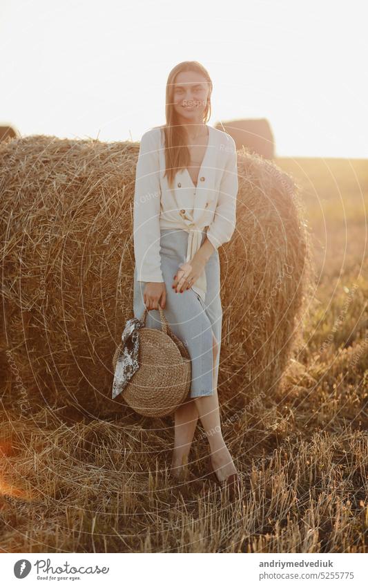Full length portrait of a smiling beautiful brunette in a jeans skirt and straw bag in hand. Woman enjoying a walk in a wheat field with hay bales on summer sunny day.