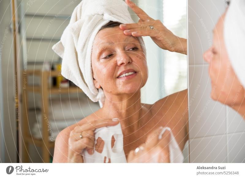 Mature beautiful woman applying green homemade DIY facial mask on skin. Looking in the mirror in bathroom, having fun. Wrapped in a towel. Skincare routine and anti-aging treatment for older women.