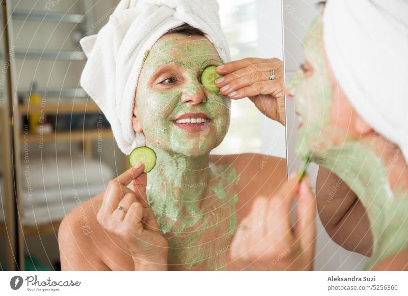 Mature beautiful woman applying green homemade DIY facial mask on skin. Looking in the mirror in bathroom, having fun. Wrapped in a towel. Skincare routine and anti-aging treatment for older women.