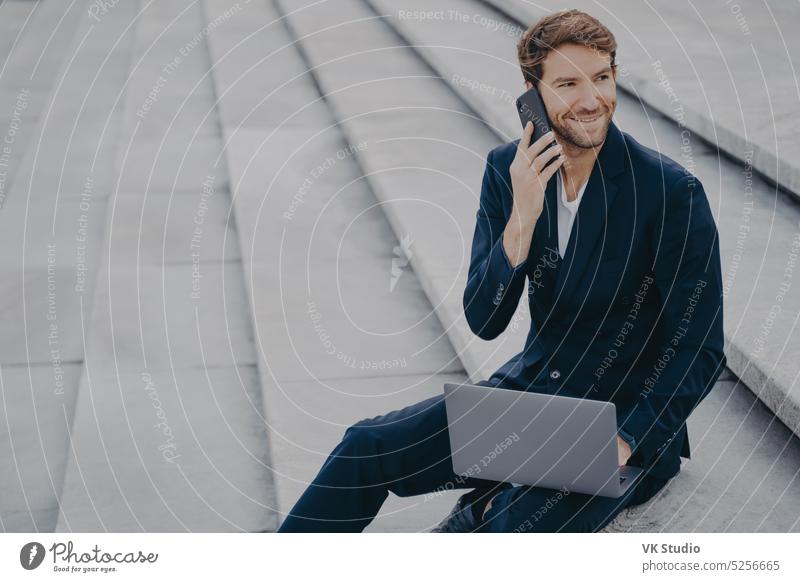 Caucasian bearded young man sitting on steps outdoor with laptop and talking on mobile phone business satisfied businessman call work remote smiling stairs